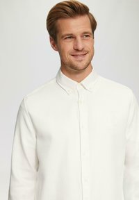 Chemise blanche à boutons en tissu texturé avec col et boutons à l'avant. La chemise a une coupe décontractée et est présentée sur un modèle souriant.