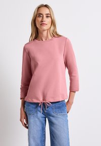 Ljusrosa tröja med rundad halsringning, trekvartslånga ärmar och dragsko neddel. Mjuk, strukturerad tyg. Matchad med jeans.