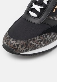 Gros plan sur le bout d'une basket noire avec des lacets blancs et une bordure en tissu marron et noir à motifs, montrant la semelle texturée et les détails de couture.