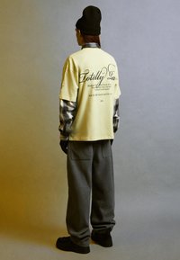 Un tricou galben supradimensionat cu text pe spate, suprapus peste o cămașă gri în carouri. Pantaloni gri largi și adidași negri completează ținuta.
