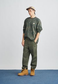 Zaļš kampa džemperis ar logotipa uzšuvumu, olīvu zaļas krāsas bikses un brūnas zābaki. Cepure ir gaiši bēša ar logotipu. Vienkāršs, vaļīgs piegriezums.