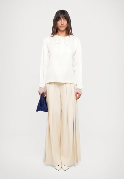 Hvit bluse med lange ermer og blonder, kombinert med beige vidde bukser. Holder en liten marineblå veske. Glatt stofftekstur.
