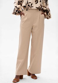 Beige wijde broek met een gladde textuur, voorzien van zijzakken en twee decoratieve knopen op de tailleband, gepaard met bruine blokhak sandalen.