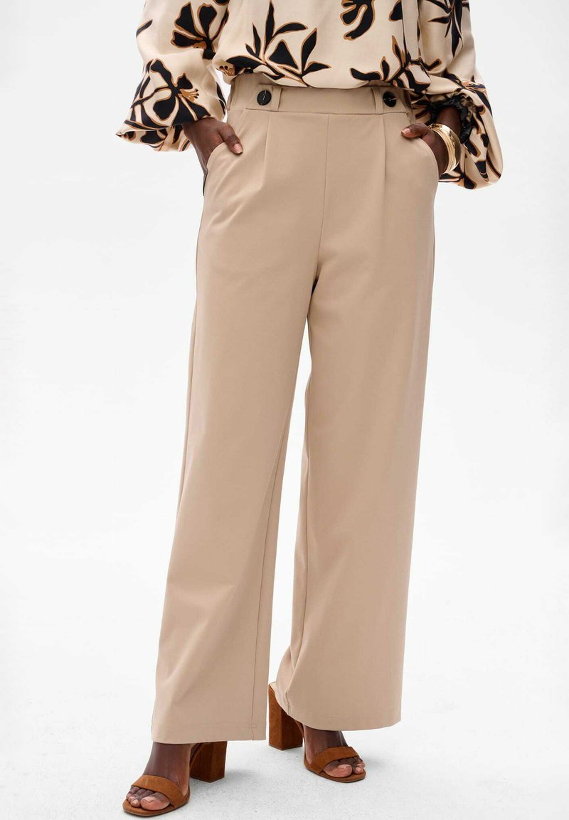 Beige wijde broek met een gladde textuur, voorzien van zijzakken en twee decoratieve knopen op de tailleband, gepaard met bruine blokhak sandalen.