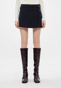 Μαύρη μίνι φούστα με επίπεδο ζώνη, λεπτομέρεια πλάγιας flap και μεταλλικό διακοσμητικό. Συνδυασμένη με ψηλές, γυαλιστερές μπότες σε καφέ χρώμα με σχέδιο ερπετού.