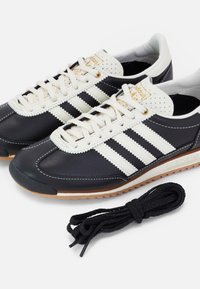 Black leather athletic shoes with white stripes and beige accents. Includes round beige laces and a black lace set. Textured rubber sole.
