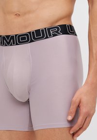 Helle lila Sportshorts mit einem schwarzen Taillengummi, auf dem der weiße Schriftzug "ARMOUR" prangt. Hergestellt aus einem glatten, dehnbaren Material mit einem enganliegenden Design.