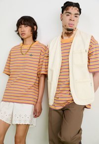 Divas modeļi valkā svītrainus T-kreklus dzeltenā, oranžā un purpursarkanā krāsā. Viens modelis ir ģērbies baltā bezpiedurkņu vestē; otrs valkā baltu svārku.