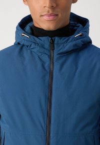 Man wearing a blue hooded jacket with a black turtleneck visible under the zipped-up front.