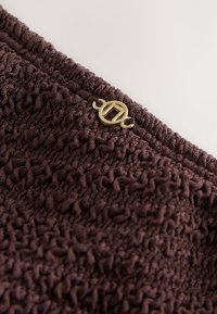 Gros plan sur un tissu texturé marron foncé avec un petit emblème métallique doré rond attaché près du bord supérieur.