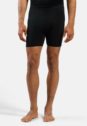 Männlich, trägt schwarze enge Shorts, steht barfuß und zeigt Tattoos an Beinen, Knöcheln und Unterarmen vor einem schlichten hellen Hintergrund.