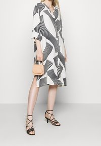 Abito bianco con strisce astratte nere, di lunghezza al ginocchio, con scollo a V e maniche a tre quarti. Tacchi neri con strappi e una piccola borsa color beige chiaro.