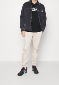 Black denim jacket with chest pockets, black graphic t-shirt, beige pants, and black sneakers with colorful accents.