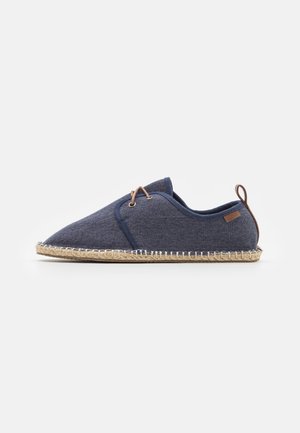 Navy blue espadrille shoe with jute sole, tan lace loop at heel, and short tan laces threaded through metal eyelets.