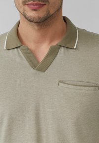 Oliwkowa koszulka polo z dekoltem w kształcie litery V, krótkimi rękawami i teksturowanym splotem. Posiada małą kieszonkę z kontrastowym wykończeniem.