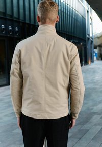 Beige, lichtgewicht jas met een hoge kraag, gestructureerde schouders en subtiele stikdetails. Gladde textuur met een aangesloten silhouet.