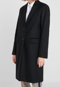 Person klädd i en svart skräddarsydd kappa över en vit skjorta, tillsammans med rutiga byxor i orange och marinblå ränder.