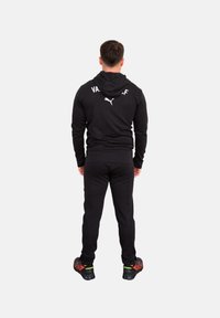 Chándal atlético negro con capucha, con un logo blanco en la parte trasera. El modelo lleva zapatillas con acentos rojos. Vista desde atrás.