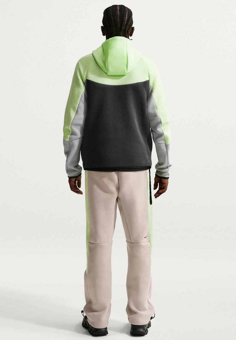 Person med flettet hår iført en hættetrøje i sort, grå og neon grøn og beige bukser med grønne striber, stående med ryggen til.