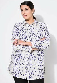 Camicia a maniche lunghe di un leggero beige con motivi a pois viola. Presenta un colletto, chiusura frontale con bottoni e spacchi laterali per facilitare l'uso.