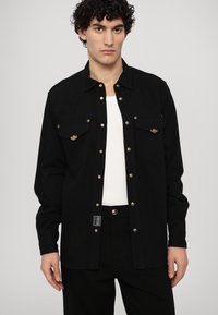 Chaqueta negra de camisa con mangas largas, botones de presión dorados, dos bolsillos en el pecho y una tela ligeramente texturizada. Usada sobre una camiseta sin mangas blanca.