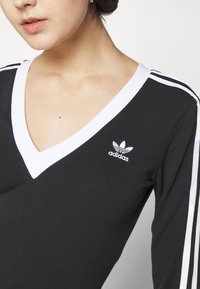 Zwarte langemouwen top met een witte V-hals kraag en witte drie-strepen detail op de mouwen; heeft een geborduurd wit Adidas-logo.