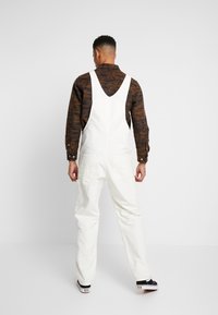 Salopette en coton blanc avec des bretelles ajustables, dotée de deux grandes poches latérales et d'une coupe droite, portée par-dessus une chemise à motifs marron.