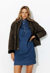 Veste en imprimé léopard marron avec des poignets noirs, portée sur une robe en denim bleu avec un devant à boutons et un col froncé.