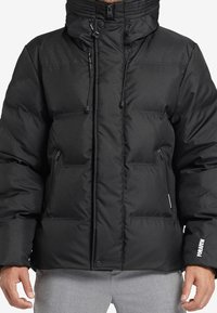 Musta toppatakki, jossa on korkea kaulus, säädettävät kiristysnyörit, vetoketjulliset etutaskut ja logokoriste hihassa. Sileä, tikattu pinta.