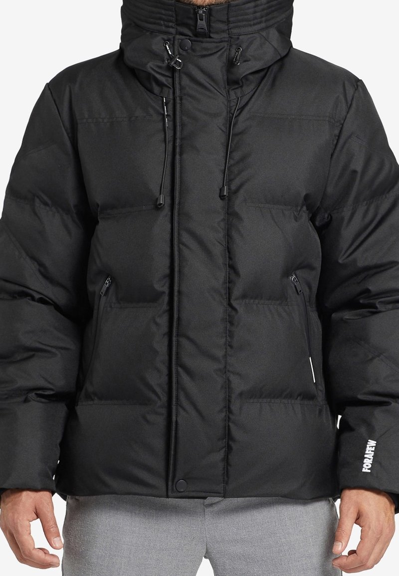 Musta toppatakki, jossa on korkea kaulus, säädettävät kiristysnyörit, vetoketjulliset etutaskut ja logokoriste hihassa. Sileä, tikattu pinta.