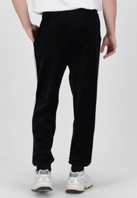 Joggers de terciopelo negro con cintura elástica y bolsillos laterales. Presenta franjas de acento blancas a lo largo de los lados. Llevados con zapatillas de color claro.