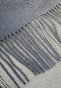 Sciarpa grigia in lana con estremità frangiate, caratterizzata da un motivo in rilievo e una texture morbida, esposta su uno sfondo neutro chiaro.