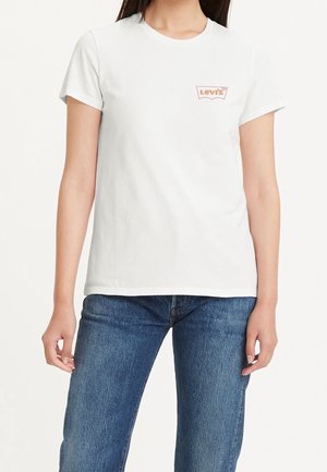 Kobieta w białej koszulce Levi's z małym logo na piersi i niebieskich dżinsach, stojąca na jednolitym tle.