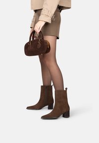 Stivaletti in camoscio marrone alla caviglia con punta affusolata, zip laterale e tacco a blocco. Accompagnati da una borsa marrone con dettagli dorati.