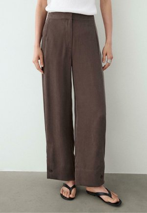 Persona che indossa pantaloni marroni a vita alta con gamba larga e dettagli con bottoni sull'orlo, una canotta bianca, infradito nere e un anello d'argento sulla mano destra.