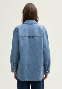 Lyseblå denimjakke med spids krave, lange ærmer og lige skæring i kanten. Har stikninger og knaplukning ved ærmerne.