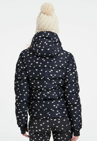 Marineblaue Pufferjacke mit weißem Blumenmuster, ausgestattet mit Kapuze und gestepptem Textil. Kombiniert mit schwarzen Hosen mit Blumenmuster.