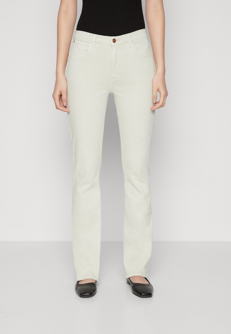 Vaqueros blancos acampanados con cinco bolsillos, trabillas para cinturón y un solo botón marrón. La tela presenta una textura suave y un acabado limpio. Se llevan zapatos planos negros.