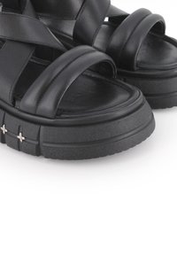 Sandalias de plataforma negras de cuero con suelas de goma gruesa y texturizada. Presenta amplias correas cruzadas y acentos florales plateados en el lateral.