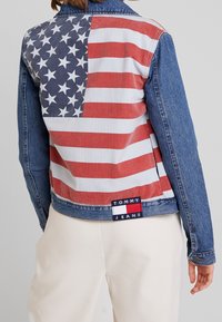 Chaqueta de mezclilla con una gran impresión de la bandera estadounidense en rojo, blanco y azul en la parte posterior, con una etiqueta de 'TOMMY JEANS' en el dobladillo.