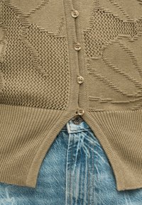 Cardigan beige en maille avec motifs texturés et quatre boutons, partiellement déboutonné sur un jean en denim bleu clair délavé à l'acide.