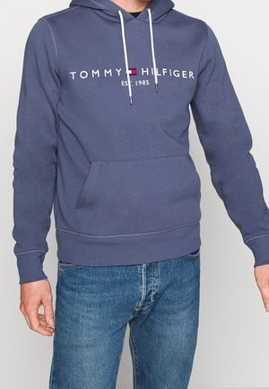 Man in een blauwe Tommy Hilfiger hoodie met een voorzak en witte trekkoorden, gecombineerd met een blauwe jeans, tegen een effen achtergrond.