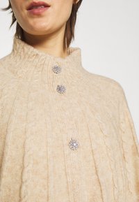 Pull en maille beige avec col haut, doté de trois boutons floraux en argent. Texture douce avec des motifs de tricot marqués.
