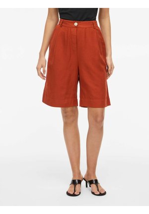 Vrouw die hooggetailleerde roestkleurige knielange shorts draagt met knopen en rits aan de voorkant, gecombineerd met zwarte sandalen met hak en een zwart topje.