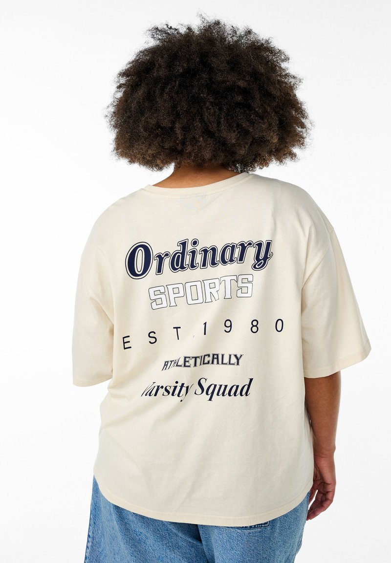 Cremefarvet oversized T-shirt med korte ærmer, der har navy blå tekst: "Ordinary Sports EST 1980 ATHLETICALLY Varsity Squad" på ryggen.