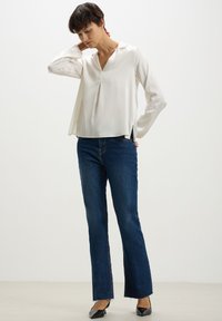 Blusa bianca a maniche lunghe con scollatura a V e spacchi laterali, abbinata a jeans svasati blu scuro. Il modello indossa ballerine nere a punta.