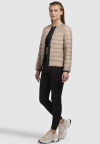 Giacca bomber imbottita in beige chiaro con colletto e polsini a coste, dotata di zip frontale e rivestimento riflettente. Indossata con leggings neri e sneakers.