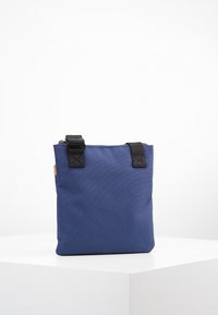 Granatowa torba na ramię wykonana z wytrzymałej tkaniny, o kwadratowym kształcie z czarnymi elementami pasków i minimalistycznym designie.