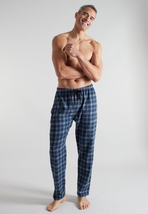 Shirtless man smiling with arms crossed, wearing blue plaid pajama pants, standing barefoot against a plain light background.