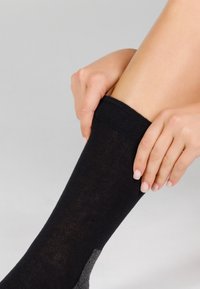 Sort sok lavet af et glat stof, med en grå detalje ved hælen. En hånd trækker sokken op, hvilket fremhæver pasformen omkring benet.